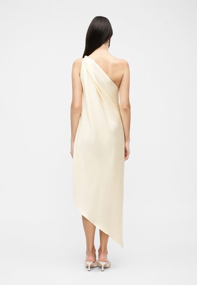 Γυναίκα με μακριά σκουρόχρωμα μαλλιά, ντυμένη με ένα κρεμ ασύμμετρο midi φόρεμα με έναν ώμο και μπεζ παπούτσια με τακούνι, στέκεται μπροστά σε απλό φόντο.