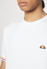 Jeune personne portant un t-shirt blanc avec un petit logo en forme de demi-cercle sur la poitrine et un détail rayé rouge et jaune sur le bord de la manche.