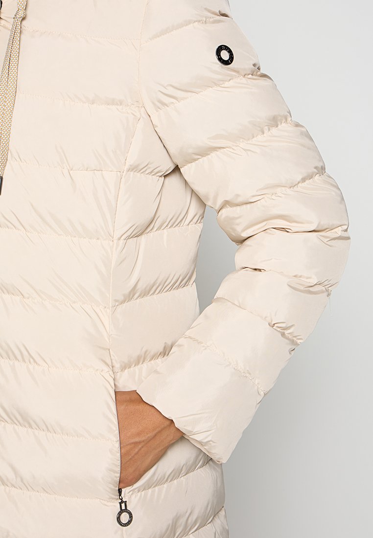 Beżowa kurtka puchowa z pikowanym wzorem, z zamkiem błyskawicznym w kieszeni oraz okrągłym logo na rękawie. Lekki materiał.