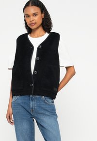 Black fleece vest with a button-up front, featuring a round neckline, side pockets, and a soft texture. Worn over a white shirt.