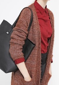 Brun stickad kofta med lös design, svart handtagsväska, kombinerad med en röd skjorta med knappar. Mjuk textur och oversized passform.