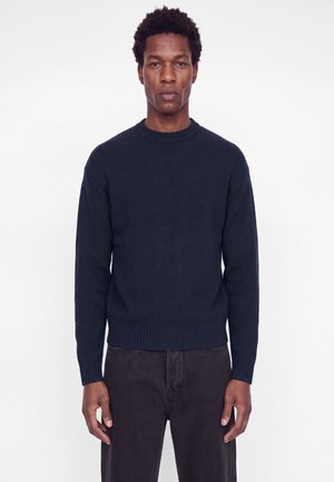 Homme aux cheveux courts et bouclés portant un pull col rond bleu marine foncé et un pantalon noir, debout sur un fond blanc uni.