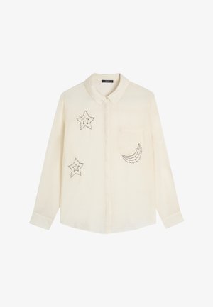 Kremowa koszula zapinana na guziki z długimi rękawami. Posiada ozdoby w postaci srebrnych gwiazdek i półksiężyca. Zawiera kieszeń z przodu.