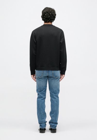 Čierna mikina s okrúhlym výstrihom a rebrovanými manžetami, v kombinácii so svetlomodrými džínsami a čiernymi topánkami. Na hlave osoby sú viditeľné kučeravé vlasy.