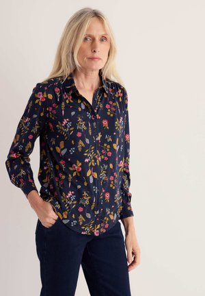 Frau mit langen blonden Haaren trägt ein dunkelblaues geblümtes Hemd und dunkle Hose, steht mit einer Hand in der Tasche vor schlichtem Hintergrund.