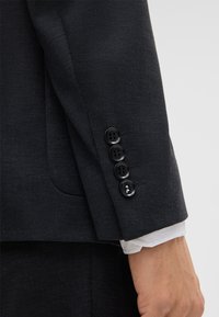 Maniche di una giacca da completo grigio scuro con quattro bottoni neri e un tessuto texture. Sotto, è visibile il polsino di una camicia bianca.