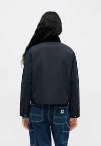 Μαύρο, κοντό μπουφάν με λεία υφή και γιακά, απεικονιζόμενο από πίσω, συνδυασμένο με μπλε φαρδιά τζιν που διαθέτουν τσέπες cargo.