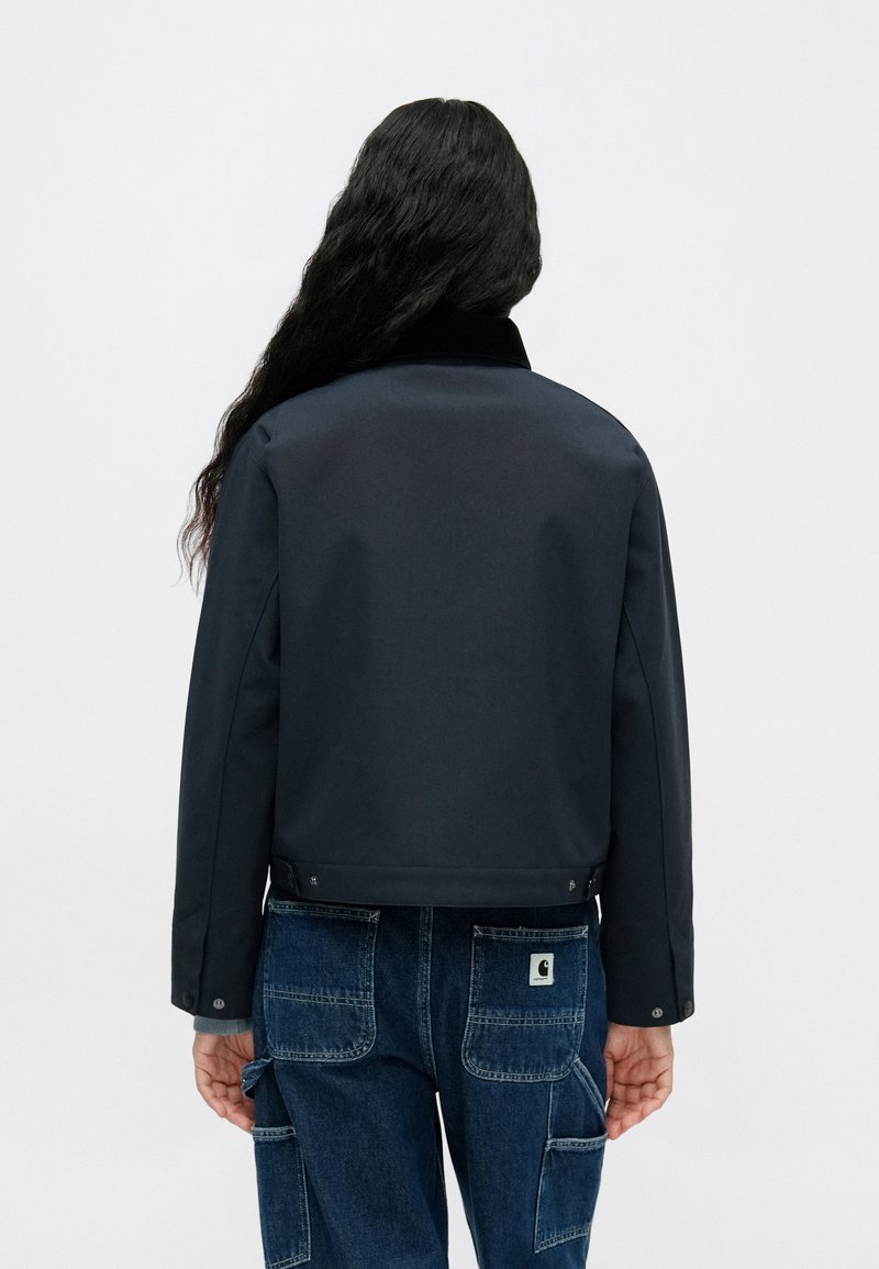 Sort, cropped jakke med en glat tekstur og ståkrave, vist fra ryggen, parret med blå baggy jeans med cargo-lommer.