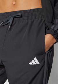 Black athletic joggers with an elastic waistband, drawstring closure, and white embroidered logo. Material is smooth and fitted with side pockets.