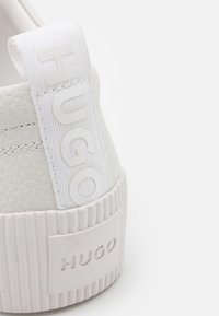 Sneaker de couro branco com acabamento texturizado, apresentando um tabuleiro de calcanhar em tecido branco com o logótipo "HUGO" em relevo e uma sola robusta com nervuras.