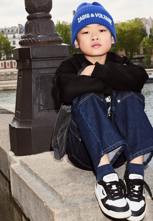 Bonnet en tricot bleu avec logo, chemise à manches longues noire, veste grise et jean bleu ample. Baskets blanches avec accents noirs. Assis près de l'eau.