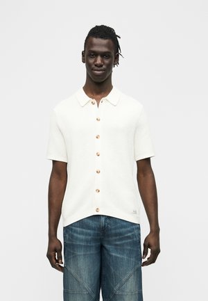 Άνδρας που φοράει λευκό πουκάμισο με κοντά μανίκια και κουμπιά και μπλε τζιν, στέκεται μπροστά σε απλό λευκό φόντο.