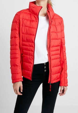 Femme portant une veste matelassée rouge vif par-dessus une chemise blanche et un pantalon noir avec trois boutons visibles à la taille.