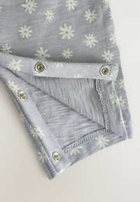 Tejido gris claro con patrón de flores blancas, que cuenta con cierres a presión plateados y un dobladillo cosido, destacando un acabado suave y texturizado.