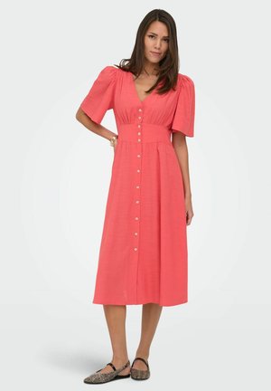 Kobieta stoi z ręką na biodrze, ubrana w koralową, zapinaną na guziki sukienkę midi z bufkami na rękawach i wzorzyste płaskie buty na jednolitym tle.