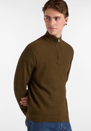 Bruine gestructureerde ritstrui met een hoge kraag en een subtiel zigzagpatroon, gecombineerd met blauwe jeans. Klein logo op de borst.