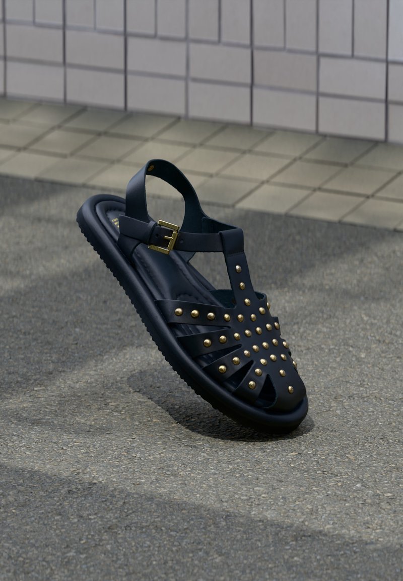 Μαύρο σανδάλι με χρυσά καρφιά και λουράκι με αγκράφα, ακουμπισμένο σε επιφάνεια από άσφαλτο κοντά σε πλακόστρωτο πεζοδρόμιο.