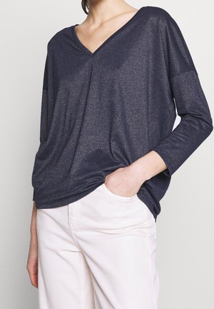Personne portant un haut ample bleu foncé à col en V et manches longues avec un pantalon blanc, une main dans une poche, sur un fond uni.