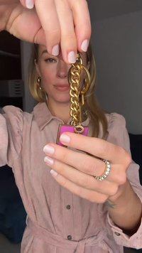 Kulcstartó arany lánccal, rózsaszín címkével és kör alakú fémalkatrésszel. A lánc vastag szemekből áll, amelyek egy téglalap alakú, színes medálhoz kapcsolódnak.