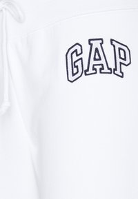 Białe spodnie dresowe z granatowym haftowanym logo "GAP" na lewym nogawce; regulowany sznurek w pasie, miękka tekstura materiału.
