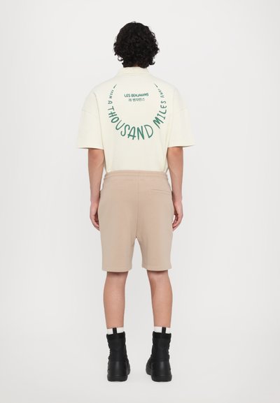 T-shirt oversize beige clair avec texte vert au dos, associé à un short beige et des bottines noires. Design simple, en matériau coton.