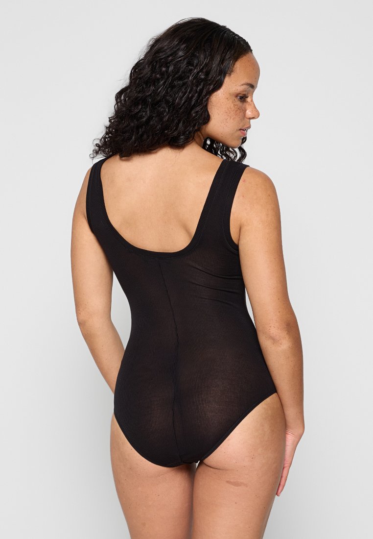 Donna con capelli ricci e scuri, che indossa un bodysuit senza maniche di colore nero, in piedi con le spalle voltate verso uno sfondo grigio chiaro uniforme.