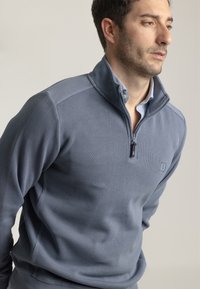 Pullover gris con cremallera hecho de tejido texturizado y con un alto cuello, que presenta un pequeño logo en el pecho y una camiseta ligera azul debajo.
