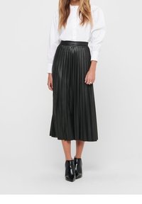 Jupe midi plissée noire en simili cuir, avec une taille haute et une texture lisse. Associée à un chemisier blanc boutonné et des bottines noires.