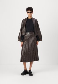 Braune Kunstleder-Bomberjacke, schwarzes Oberteil und brauer Faltenrock mit Schlangenmuster. Schwarze Schuhe runden das Outfit ab. Sonnenbrille getragen.