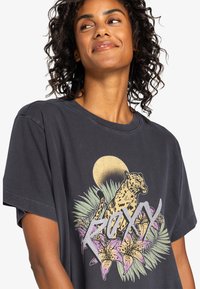 T-shirt in cotone grigio scuro con grafica di una leopardessa, circondata da foglie verdi e gigli rosa, con il testo "ROXY" in argento. Maniche corte.