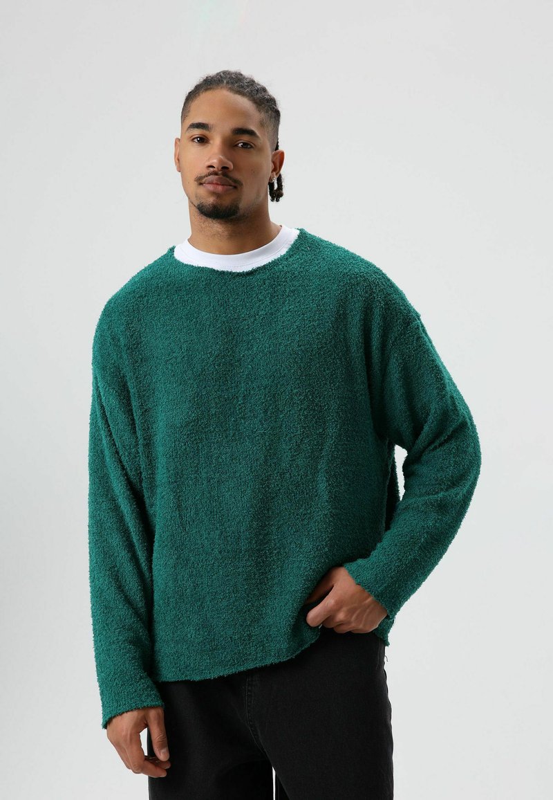 Oversized groene gebreide trui met lange mouwen, ronde halslijn en ruwe randen, gedragen over een wit shirt. Zwarte jeans zichtbaar.