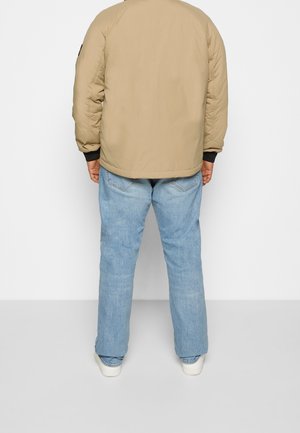 Osoba ubrana w beżową kurtkę, jasnoniebieskie dżinsy i białe trampki, stojąca tyłem na jednolitym białym tle.