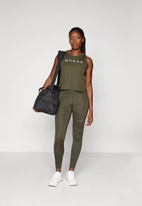 Oliwkowy zestaw sportowy składający się z bezrękawnika i legginsów z subtelnym logo. Modelka trzyma czarną torbę na siłownię, nosząc białe sneakersy.