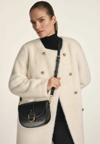 Czarna skórzana torba na ramię z złotymi akcentami, o zaokrąglonym kształcie i z klapką. Wełniany płaszcz w odcieniu kości słoniowej z złotymi guzikami i fakturowaną powierzchnią.