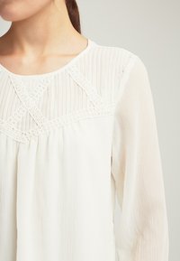 Bílá blůza s vertikálními sklady, lodičkovým výstřihem a složitými krajkovými detaily na těle. Dlouhé, splývavé rukávy zvýrazňují design.