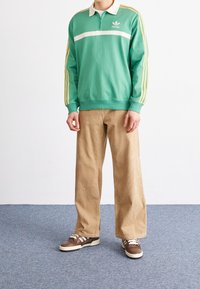 Grön tröja med vit rand och dragkedja vid kragen, parat med beiga kordbyxor och bruna sneakers med vita detaljer.