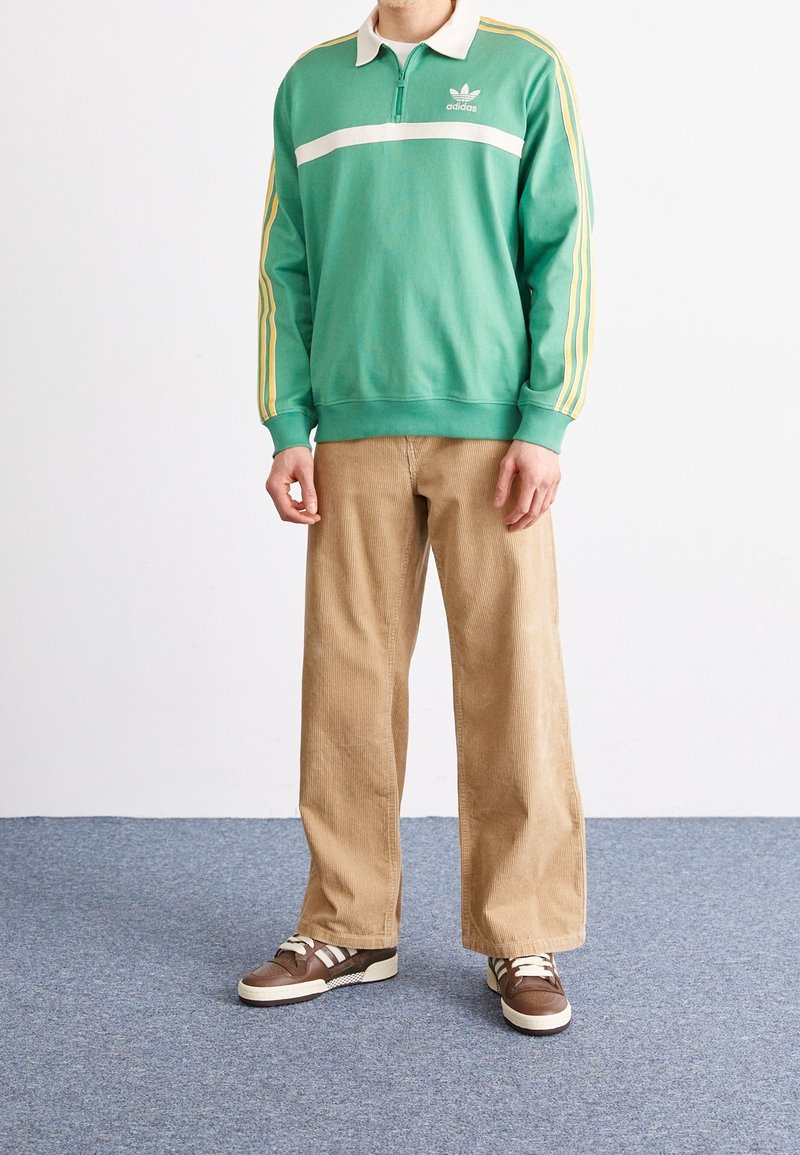 Grön tröja med vit rand och dragkedja vid kragen, parat med beiga kordbyxor och bruna sneakers med vita detaljer.