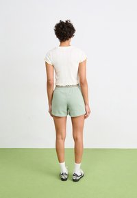 Bílý elastický crop top s žlutým lemem na rukávech, doplněný zelenými šortkami. Model má na sobě bílé ponožky a černé boty s puntíkovaným vzorem.