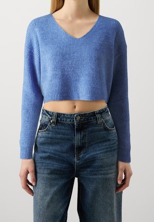 Woman wearing a cropped blue knit sweater with long sleeves and high-waisted dark blue jeans, standing against a plain background.