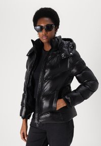 Black puffer jacket with shiny finish, zip closure, and detachable hood; features side pockets and elastic cuffs. Worn with black top and sunglasses.