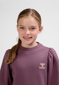 Lila Sweatshirt mit Puffärmeln, geripptem Halsausschnitt und kleinem, gesticktem Logo auf der Brust. Glatte Stoffstruktur.