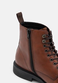 Smeđe kožne čizme do gležnja s bočnim patent zatvaračem, vezicama i teksturiranim potplatom. Imaju petlju za lakše obuvanje. Glatka, izdržljiva završna obrada.