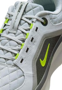 Gros plan d'une chaussure Nike gris clair avec languette matelassée, boucles de lacets vertes et virgule Nike vert néon sur un panneau latéral texturé foncé.