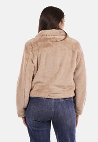 Giacca in pelliccia sintetica beige con maniche lunghe, colletto e vestibilità aderente, progettata con una texture liscia e minimi dettagli sulla parte posteriore.