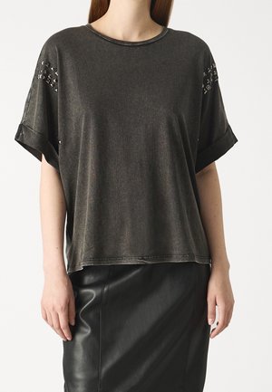 Mujer con una blusa suelta de manga corta de color gris oscuro con adornos de tachuelas en las mangas y una falda de cuero negra, de pie frente a un fondo blanco.