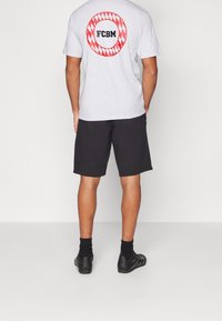 Muž má na sobě světle šedé tričko s červeným a bílým kruhem a textem "FCBM" na zádech, černé kraťasy, černé ponožky a černé sportovní boty.