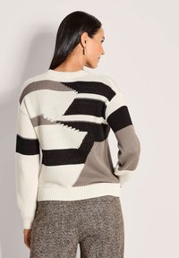 Vrouw in een gebreide trui met geometrische patronen in zwart, wit en grijs, gecombineerd met getextureerde bruine broek, staande opzij.