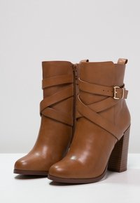 Hnědé kožené kotníkové boty s vysokým blokovým podpatkem, zdobené překříženými pásky a bočním zipem. Hladká textura s lesklým povrchem.