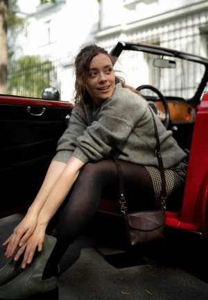 Red vintage car with black interior. Person in grey sweater, black tights, and olive green ankle boots, leaning out of the door. Brown handbag.