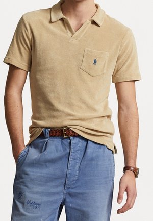 Mężczyzna w beżowej koszulce polo z krótkim rękawem i logo na kieszeni piersiowej, w niebieskich zniszczonych spodniach, z brązowym plecionym paskiem i skórzanym zegarkiem na nadgarstku.